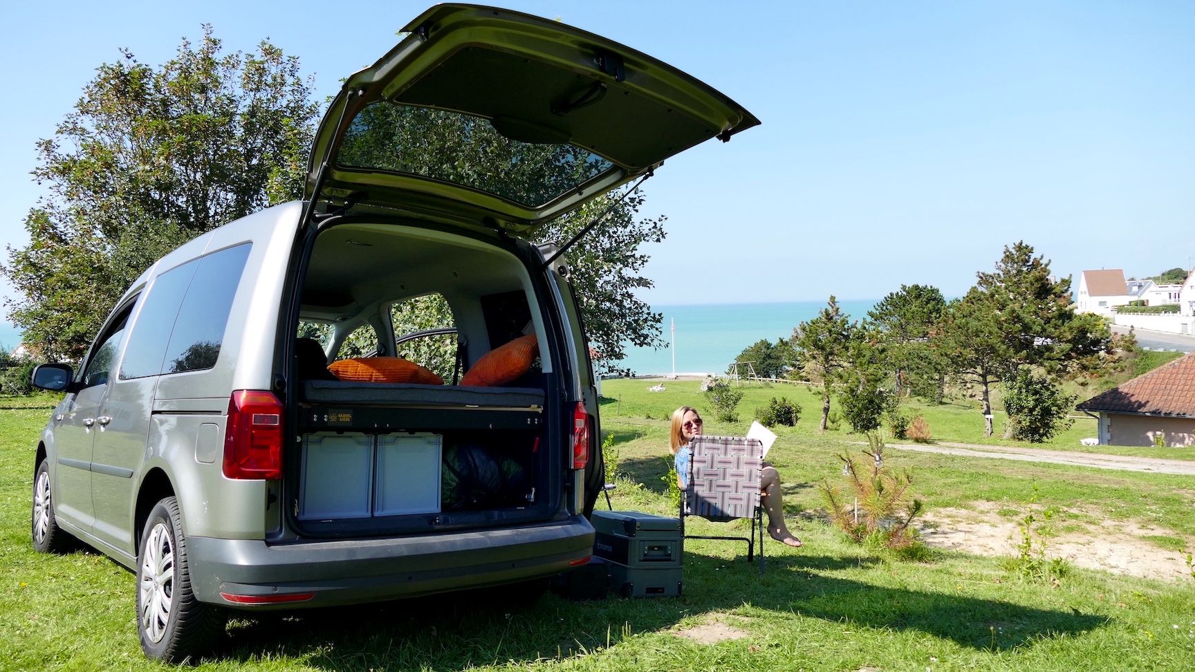 VW Caddy mit offenem Kofferraum auf einem Campingplatz, Frau sitzt im Stuhl und liest.