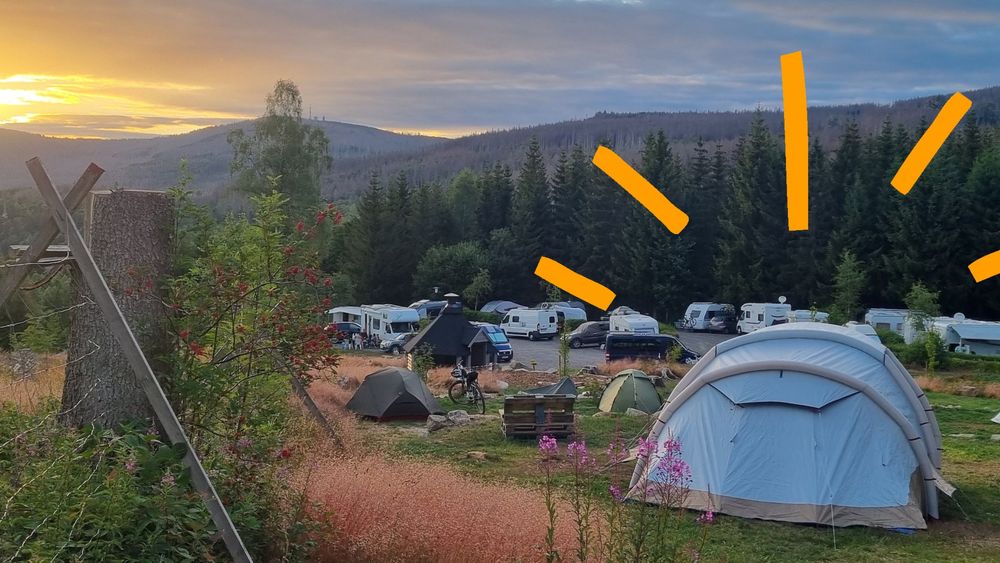 Zelt und Campingfahrzeuge auf einer Wiese mit Blick auf Wälder im Sonnenuntergang.