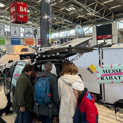 Well-attended exhibition stand at the camping trade fair. Visitors stand around the VW Caddy and look at the camping bed.