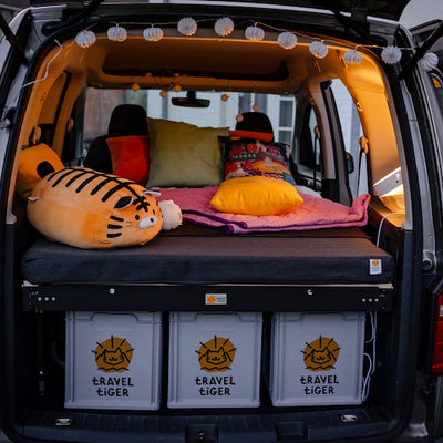 VW Caddy steht mit offenem Kofferaum und Campingbett auf einer Wiese.